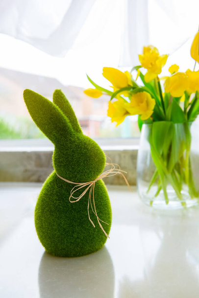 Cute green grass bunny rabbit and bouquet of fresh spring yellow tulips and daffodils flowers on the white kitchen table near the window. Happy Easter vertical card. Selective focus. Copy space. - Foto, imagen