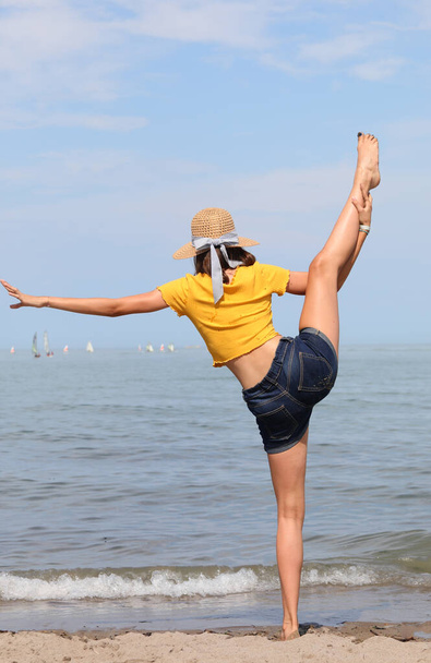 junges schlankes Mädchen turnt im Sommer am Meer und hebt das Bein mit dem Fuß über dem Kopf - Foto, Bild