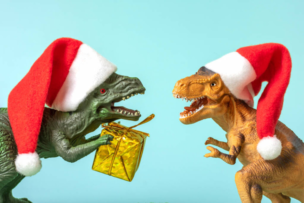 赤いサンタクロースの帽子の2つの恐竜レックスピンクの背景にその爪で黄金のギフトボックスを保持大晦日やクリスマスイブアートホリデーカードメリークリスマスのコンセプトのための創造的なアイデア. - 写真・画像