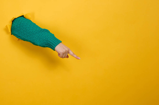 la mano femenina sobresale de un agujero roto en un fondo de papel amarillo, parte del cuerpo apunta con el dedo índice hacia un lado. Copiar espacio - Foto, Imagen