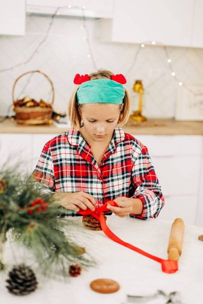 Ένα όμορφο κορίτσι διακοσμεί τα μπισκότα με μια φωτεινή κόκκινη κορδέλα, προετοιμάζει ένα γλυκό κέρασμα για τους επισκέπτες τα Χριστούγεννα ή την παραμονή της Πρωτοχρονιάς - Φωτογραφία, εικόνα