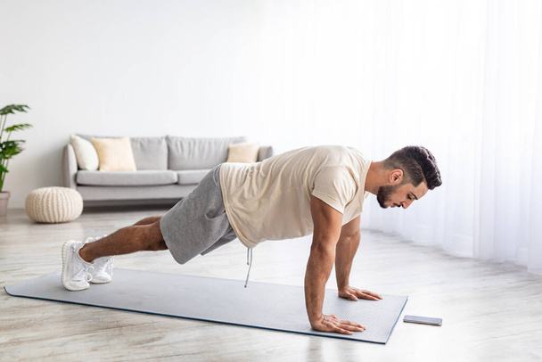Handsome young Arab man doing plank, following online video tutorial on smartphone at home, full length - Fotó, kép