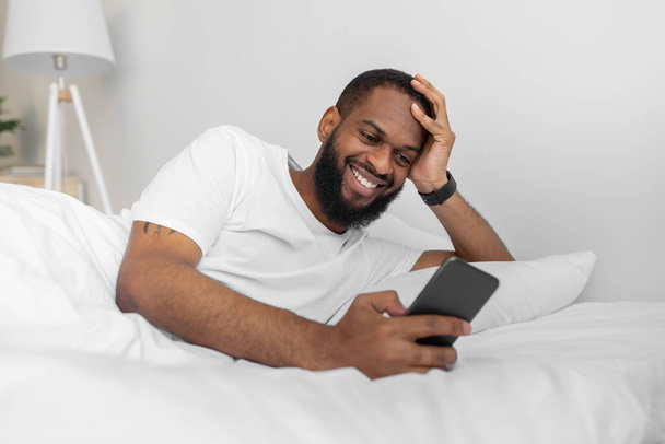 Ευτυχισμένος νεαρός αφροαμερικάνος άντρας με γενειάδα δακτυλογραφεί στο τηλέφωνο και βλέποντας βίντεο στο λευκό κρεβάτι στην κρεβατοκάμαρα - Φωτογραφία, εικόνα