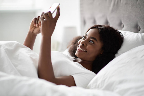 Cheerful young black woman chatting with boyfriend in bed - 写真・画像