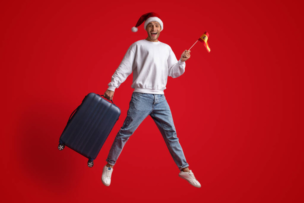 Hombre feliz viajero con sombrero de Santa saltar con bandera española y maleta - Foto, imagen