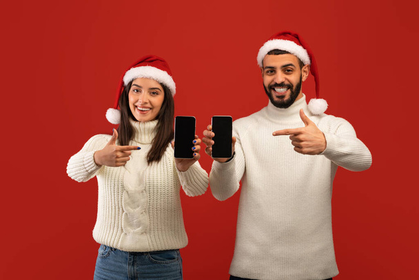 Weihnachtswerbung. Arabische Ehepartner zeigen mit dem Finger auf Handybildschirme und tragen Nikolausmützen über rotem Hintergrund - Foto, Bild