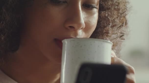 Κοντινό πλάνο πορτρέτο αγνώριστης αφροαμερικάνας γυναίκας που πίνει τσάι ή καφέ και σερφάρει στο διαδίκτυο στο κινητό - Πλάνα, βίντεο