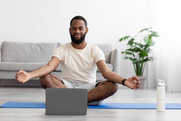 Calme jeune homme afro-américain avec barbe en vêtements de sport blancs avec les yeux fermés pratiquant le yoga en position lotus - Photo, image