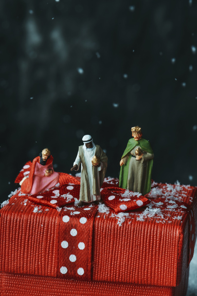 closeup of the miniatures of the magi on top of a red gift box tied with a satin red ribbon patterned with white dots, on a dark background with some blank space on top - Photo, Image
