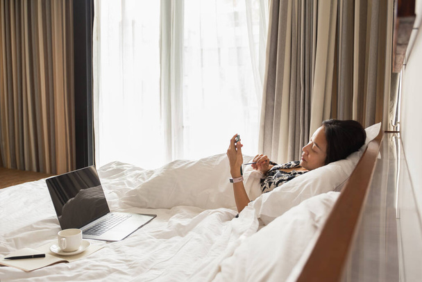 Woman using cell phone and laptop in bed on vacation. - Fotoğraf, Görsel