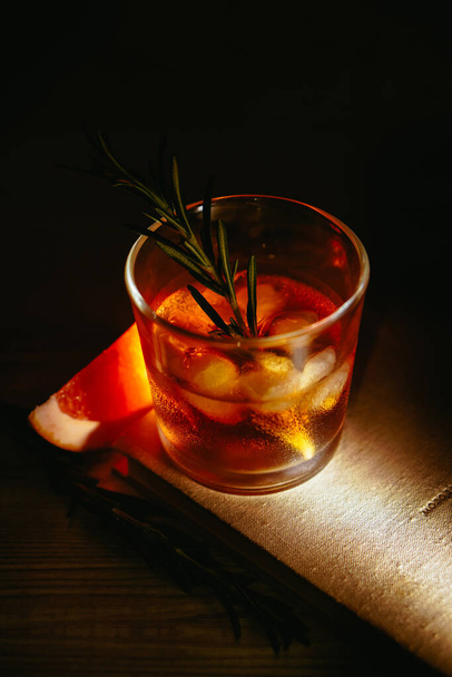 Whiskey sour cocktail with rum ice grapefruit and rosemary in shot glass on book. Alcohol cocktail in light beam - Foto, Imagem
