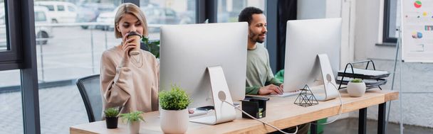 blonde woman drinking coffee to go and looking at computer monitor near african american colleague, banner - Foto, Imagen