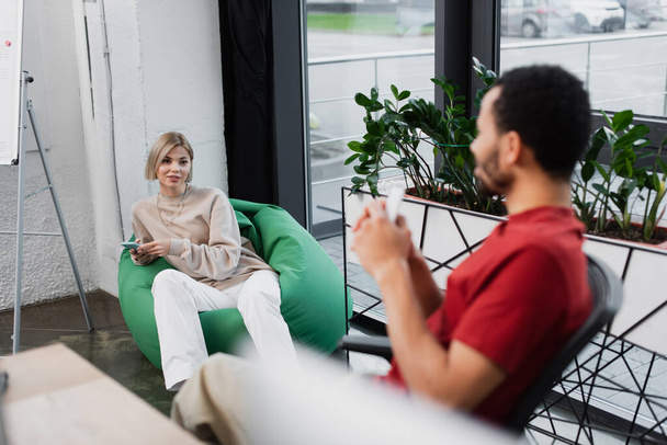 blonde manager sitting in bean bag chair and holding smartphone near blurred african american colleague in office  - Foto, Bild