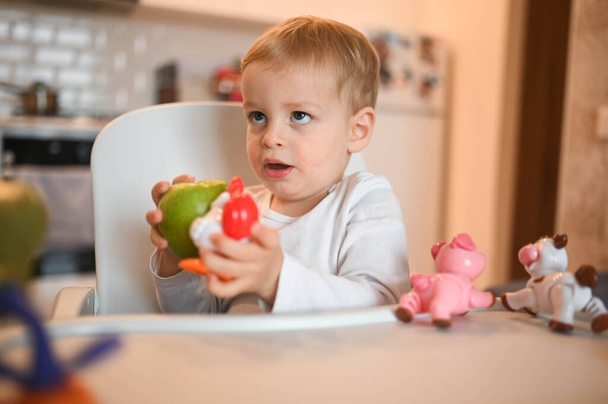 小さな幸せなかわいい赤ちゃんの幼児の男の子のブロンドは、赤ちゃんの椅子に座ってリンゴで遊んでいます。自宅のキッチンのインテリアで食べ物と赤ちゃんの顔の表情。健康的な食事幸せな家族の子供時代のコンセプト. - 写真・画像