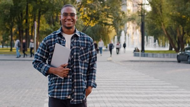 jovem afro-americano bem sucedido cara vestindo óculos de sol homem de negócios masculino com laptop sorrindo stands contra crosswalk estrada tráfego fundo olhando para câmera posando na rua sorrisos amigável - Foto, Imagem