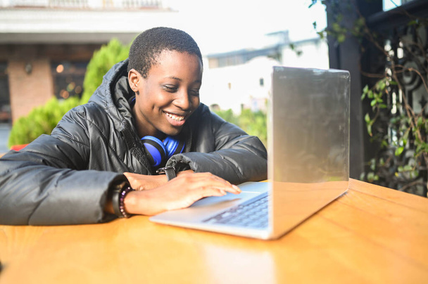 Smiling happy African black short-haired woman student in black down jacket with blue headphones studying online working on laptop computer at street cafe. Diversity. Remote work, distance education. - Photo, Image