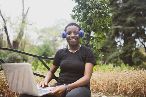 Sorrindo feliz Africano preto de cabelos curtos mulher estudante afro cabelo com fones de ouvido azuis estudando on-line trabalhando no computador portátil no verão parque verde. Diversidade. Trabalho à distância, educação à distância. - Foto, Imagem