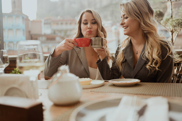 Крупный план портрета двух подруг в строгих костюмах, смеющихся, пьющих кофе и вино на террасе на летней улице кафе на заднем плане здания Старого Тбилиси, Грузия - Фото, изображение
