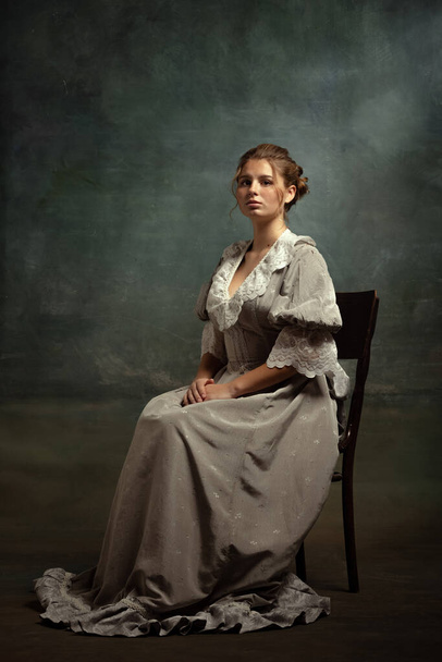 Sad young beautiful girl in gray dress of medieval style sitting on chair isolated on dark vintage background. Comparison of eras concept, flemish style. - Photo, Image