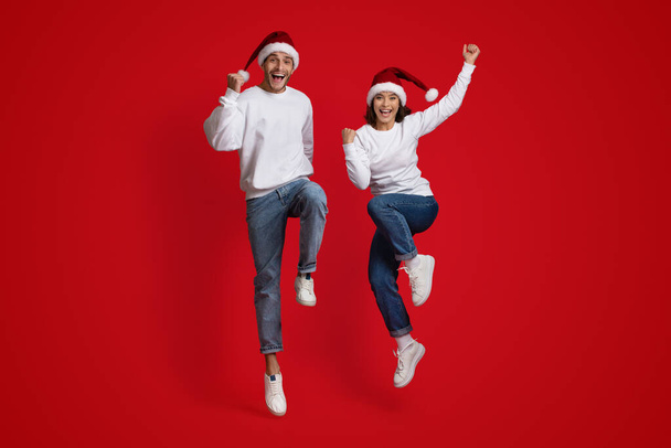 Feiertagsstimmung. Überglückliches Paar mit Weihnachtsmütze springt auf rotem Studiohintergrund - Foto, Bild