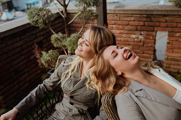 Zbliżenie portret dwóch przyjaciółek w surowych szarych garniturach śmiejących się siedząc na wiklinowych krzesłach na tarasie na zewnątrz w letniej kawiarni ulicznej. - Zdjęcie, obraz