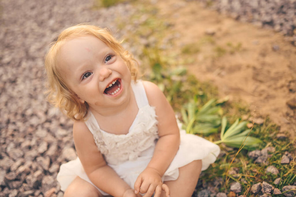 Little funny cute blonde girl child toddler with curls in white dress and with mud on her face sits on the ground on the street outside at summer. Baby laughing. Happy healthy childhood concept. - Foto, imagen