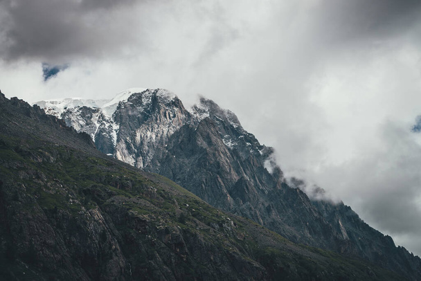 Donker atmosferisch surrealistisch landschap met donkere rotsachtige bergtop in lage wolken in grijze bewolkte lucht. Grijze lage wolk op hoge top. Hoge zwarte rots met sneeuw in lage wolken. Surrealistische sombere bergen. - Foto, afbeelding