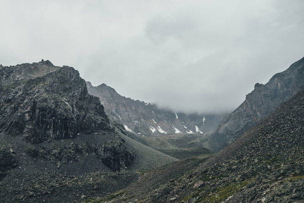 Donker atmosferisch berglandschap met grijze lage wolken in donkere vallei op grote hoogte bij regenachtig weer. Grote rotsachtige bergwand met sneeuw in lage wolken in grijze bewolkte lucht. Gloomy berglandschap - Foto, afbeelding