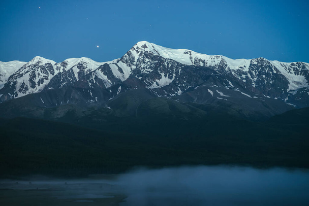 Atmospheric landscape with dense fog on lake and great snowy mountain ridge under night sky. Alpine scenery with thick fog on mountain lake and big mountain range in night. Snow pinnacle in dusk. - Photo, Image