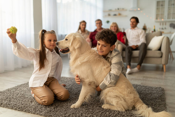 Cute children playing with their golden retriever on floor at home, their relatives sitting on couch on background - Photo, Image