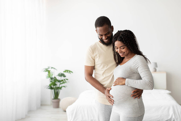 Sorridente giovane marito afroamericano abbraccia moglie incinta, toccando pancia in camera da letto minimalista interno - Foto, immagini