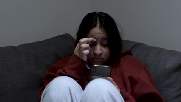 Close up shot of a young girl sitting on a couch and checking her phone. She is scratching her forehead and playing with her hair while checking phone - Filmati, video