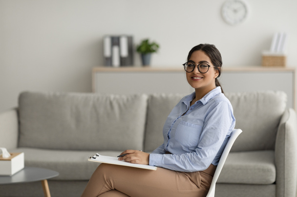 Zelfverzekerde arabische vrouwelijke psycholoog schrijft in klembord, zit op modern kantoor en glimlacht naar de camera, vrije ruimte - Foto, afbeelding