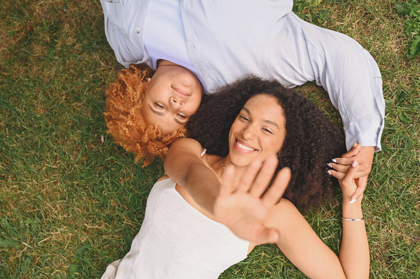 Mladý krásný šťastný lesbický afroamerický pár ležící na trávě a smějící se natahující se ruce z fotoaparátu, letní pohled shora. Komunitní koncept LGBT. Ženy přátelé těší milostné okamžiky. - Fotografie, Obrázek
