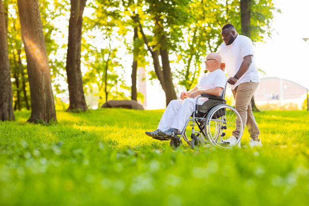 Caregiver and old man in a wheelchair. Professional nurse and patient walking outdoor in the park at sunset. Assistance, rehabilitation and health care. - Фото, изображение