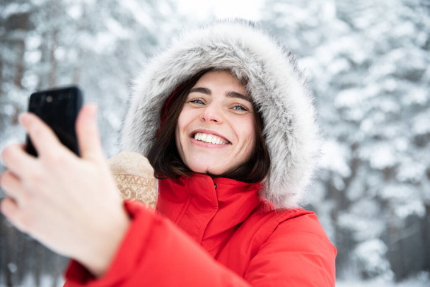 Portrait of beautiful young laughing brunette with gray eyes in red jacket with hood made of faux fur on winter day against background of snowy trees Woman takes selfie on her mobile phone and talks - Fotografie, Obrázek