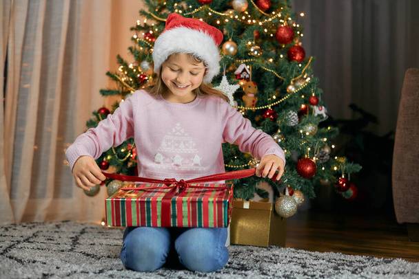Ένα κορίτσι ανοίγει ένα δώρο για την Πρωτοχρονιά δίπλα σε ένα δέντρο της Πρωτοχρονιάς. Καλά Χριστούγεννα και ευτυχισμένο το νέο έτος - Φωτογραφία, εικόνα