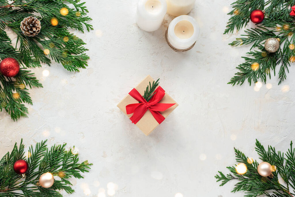 Kırmızı kurdeleyle süslenmiş Noel hediyesi kutusu. Beyaz desenli arka planda dallar, mumlar ve Noel topları. Üst manzara, düz uzanma. - Fotoğraf, Görsel