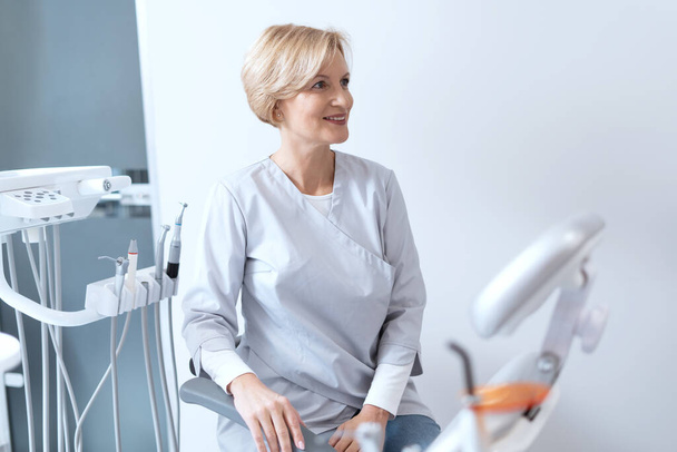 Muotokuva hymyilevä hammaslääkäri istuu lähellä laitteita ja katselee pois. Stomatologian klinikan henkilökunta. Suullinen lääketiede käsite. Hampaiden sairauksien ehkäisy - Valokuva, kuva
