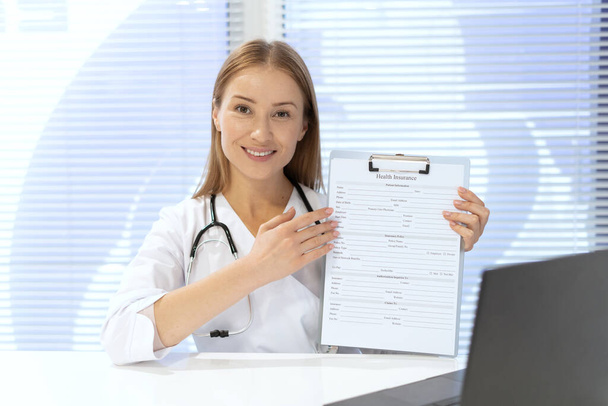 Portrait of smiling doctor with stethoscope and health insurance document. General practitioner in contemporary hospital office. Healthcare concept. Medical staff - Foto, afbeelding