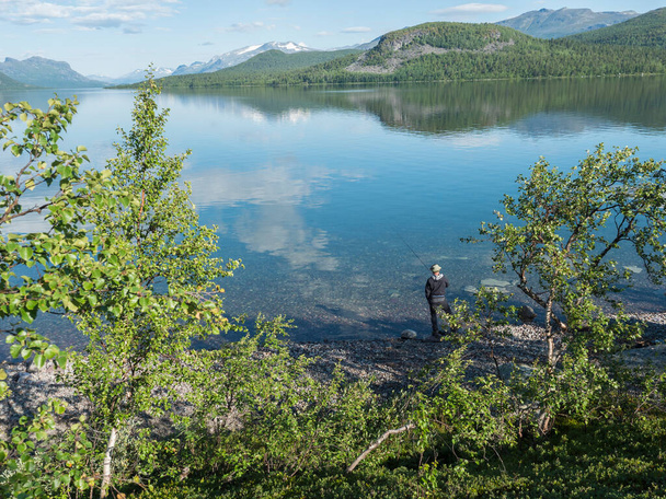 ルール川の漁師、晴れた夏の日にスウェーデンのSaltoluoktaの澄んだ穏やかなLulealvenダム湖の淡水。緑の岩の山、白樺の木や雲. - 写真・画像