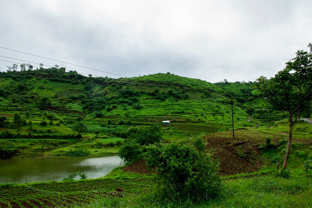 Fotografia Stockowa malowniczego widoku na zbocza pasma górskiego pokryte zielenią gruntów uprawnych i mały domek lub chata znajduje się w środku pasma górskiego.Jezioro wodne płynące wokół góry - Zdjęcie, obraz