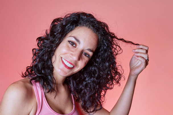Portrait of beautiful young woman with black curly hair holding one of her curls with her hand looking at camera over pink background. hair care concept. curly girl method - 写真・画像