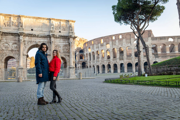Νεαρό ζευγάρι ταξιδεύει στη Ρώμη. Το νεαρό ζευγάρι ποζάρει μπροστά από την αψίδα του Τίτου. Η ξανθιά με το κόκκινο μπουφάν βάζει το χέρι της στο αγόρι της.. - Φωτογραφία, εικόνα