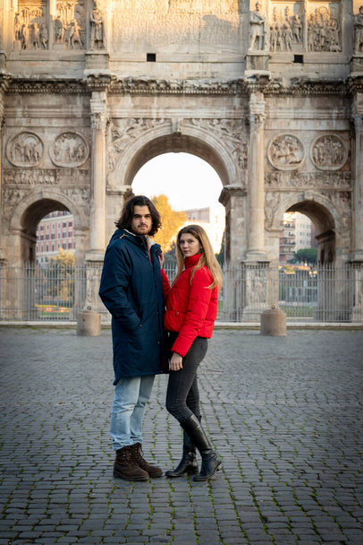 Junges Paar auf dem Weg nach Rom. Das junge Paar posiert vor dem Titusbogen. Die blonde Frau in ihrer roten Jacke legt ihre Hand auf ihren Freund. - Foto, Bild
