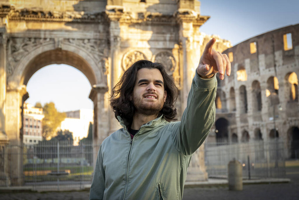 Onnellinen hetki matkalla Roomaan. Nuori hymyilevä mies poseeraa kuvassa Tituksen kaaren edessä. Pitkätukkainen mies osoittaa sormella oikeaan suuntaan. - Valokuva, kuva