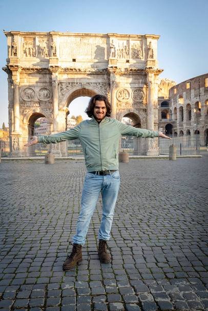 Счастливый момент в Риме. Молодой улыбающийся мужчина позирует для фотографии, раскидывая руки перед аркой Тита. - Фото, изображение