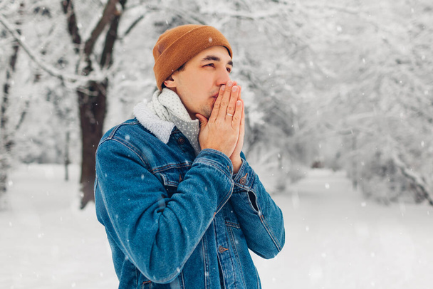 Портрет молодої людини в засніженому зимовому парку в джинсовій куртці, капелюх відчуває холодний вихід на вулицю. Хлопець зігріває руки диханням
 - Фото, зображення