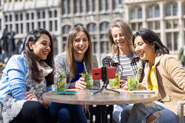 Antwerp, Belçika, 21 Mayıs 2021, dört farklı ırktan bayan turist veya öğrenci eski şehir merkezindeki bir terasta cep telefonuyla video görüşmesi yapıyorlar. - Fotoğraf, Görsel