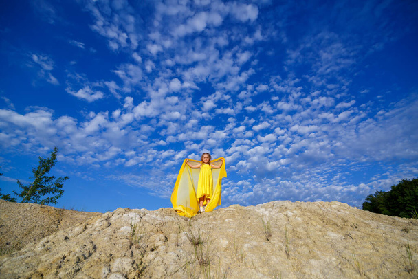 A blonde girl of 7-8 years old in a bright yellow dress against a blue sky. - Foto, afbeelding